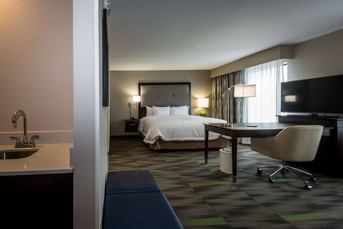 A hotel room with a bed, bedside table, lamp, desk, chair, TV, and a sink.