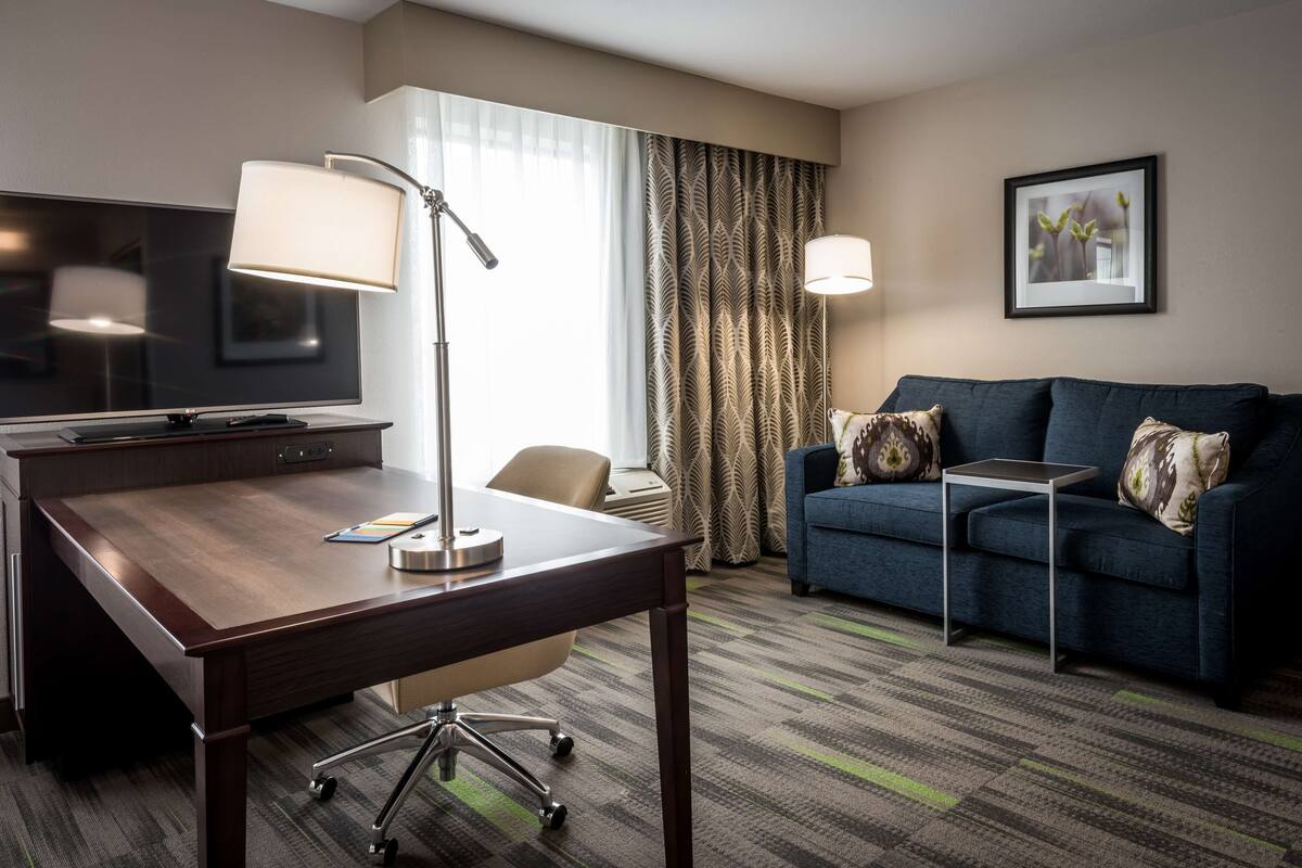 A hotel room with a desk, chair, sofa, and a TV.