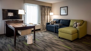 A hotel room with a desk, chair, television, sofa, and ottoman.