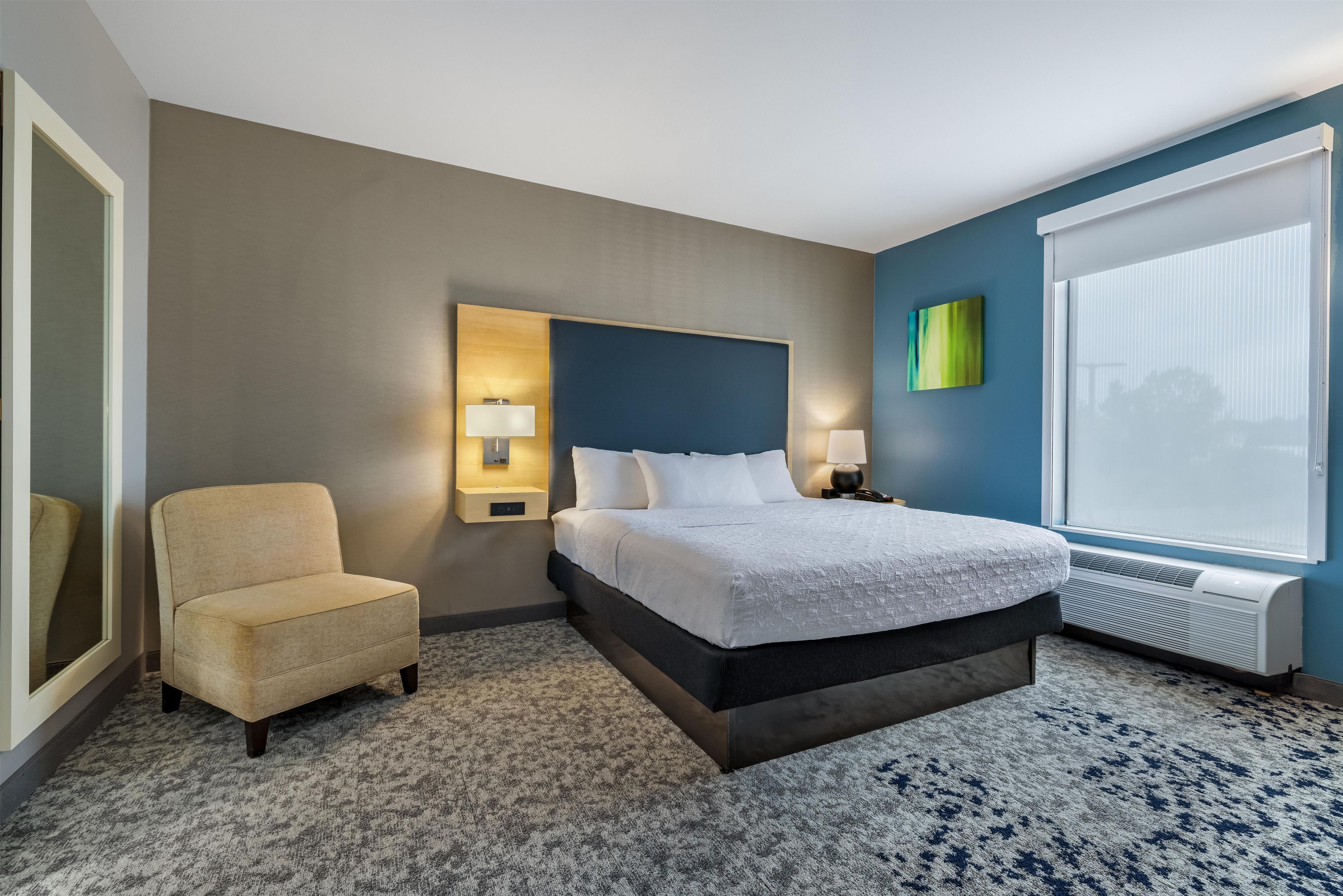 Una habitación de hotel con cama, una silla, una mesita de noche, una lámpara de pared y una ventana con persianas.
