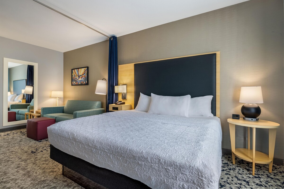A hotel room with a large bed, two bedside tables with lamps, a sofa, a red ottoman, a mirror, and a painting on the wall.