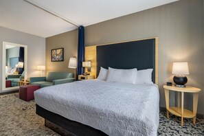 Quarto de hotel com uma cama grande, duas mesas de cabeceira com luminárias, um sofá, um banquinho vermelho, um espelho e um quadro na parede.