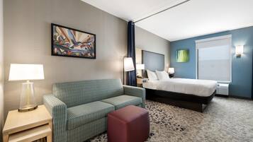 A hotel room with a bed, a sofa, a small table with a lamp, a painting on the wall, and a window with blinds.
