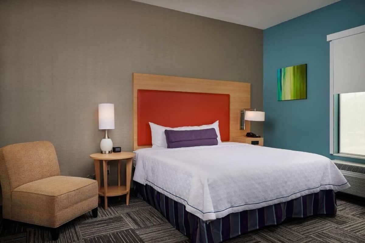 Habitació d'hotel amb un llit, una cadira, una tauleta de nit, una làmpada i una finestra.