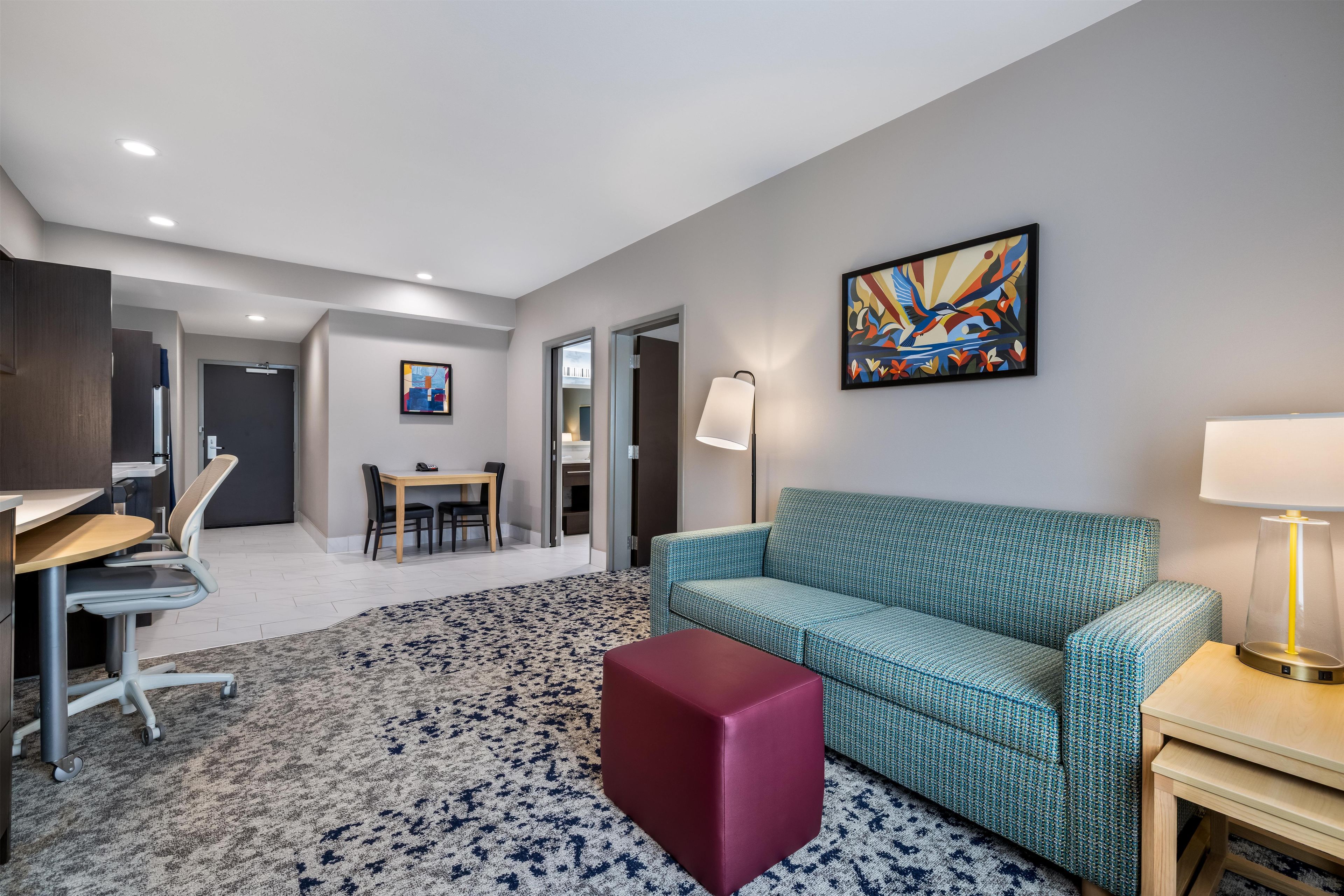 Quarto de hotel moderno com um sofá azul-esverdeado, um banquinho vermelho, uma área de refeições com mesa e cadeiras, um quadro na parede e um abajur em uma mesinha de cabeceira.