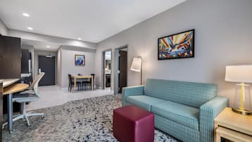 Quarto de hotel moderno com um sofá azul-esverdeado, um banquinho vermelho, uma área de refeições com mesa e cadeiras, um quadro na parede e um abajur em uma mesinha de cabeceira.