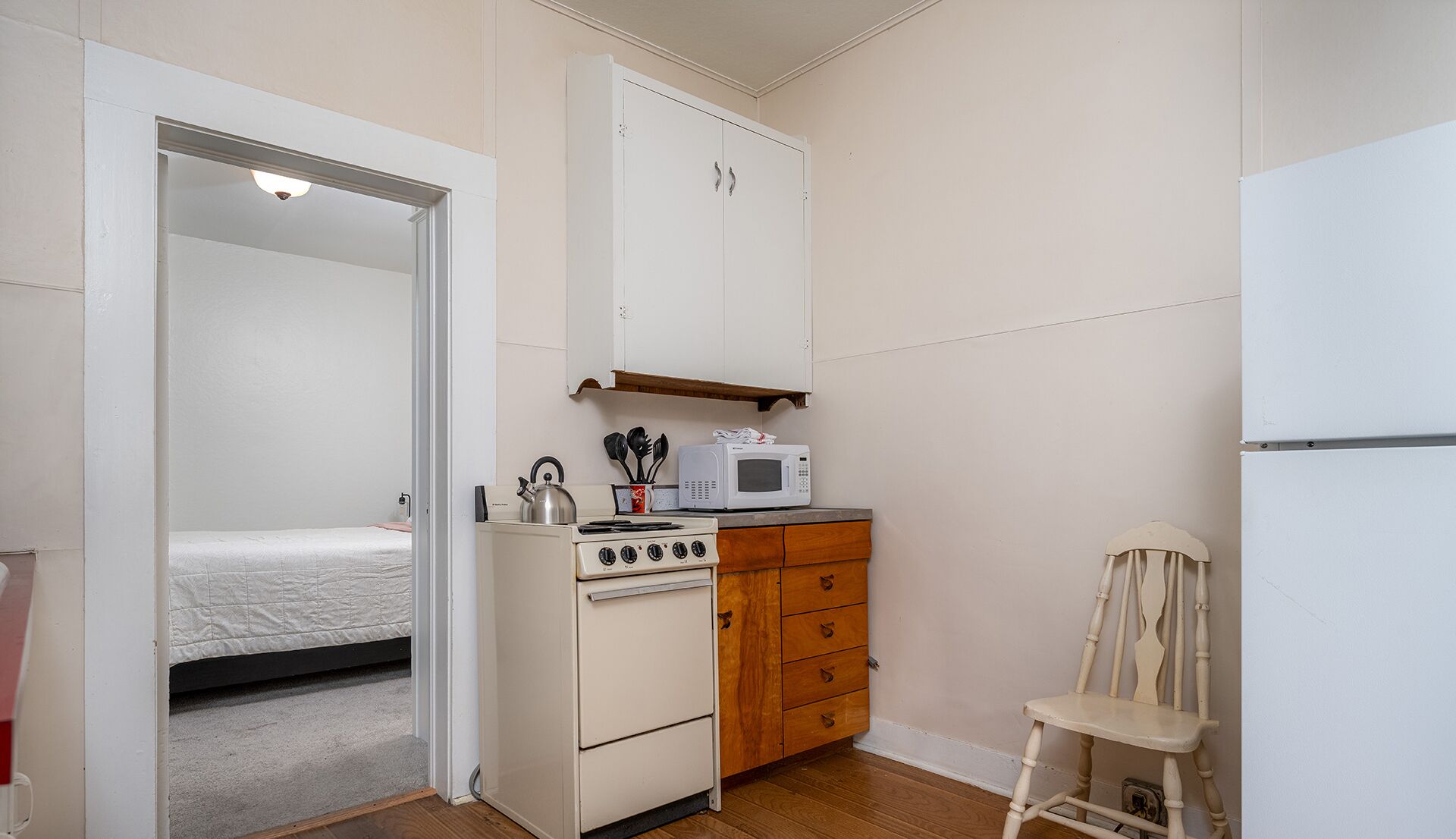 Uma cozinha compacta com fogão, micro-ondas e armários de madeira. Uma cama é visível no quarto vizinho.