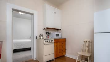 Uma cozinha compacta com fogão, micro-ondas e armários de madeira. Uma cama é visível no quarto vizinho.