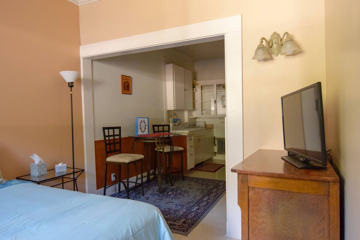 A room with a bed, a desk, a chair, a television, and a kitchenette.