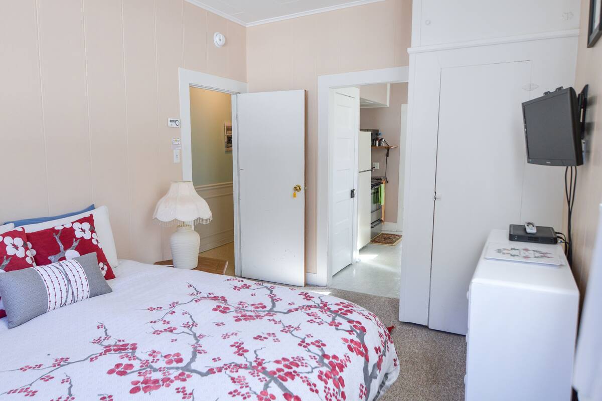 A bedroom with a bed, a white wardrobe, a television, and a door leading to a hallway.