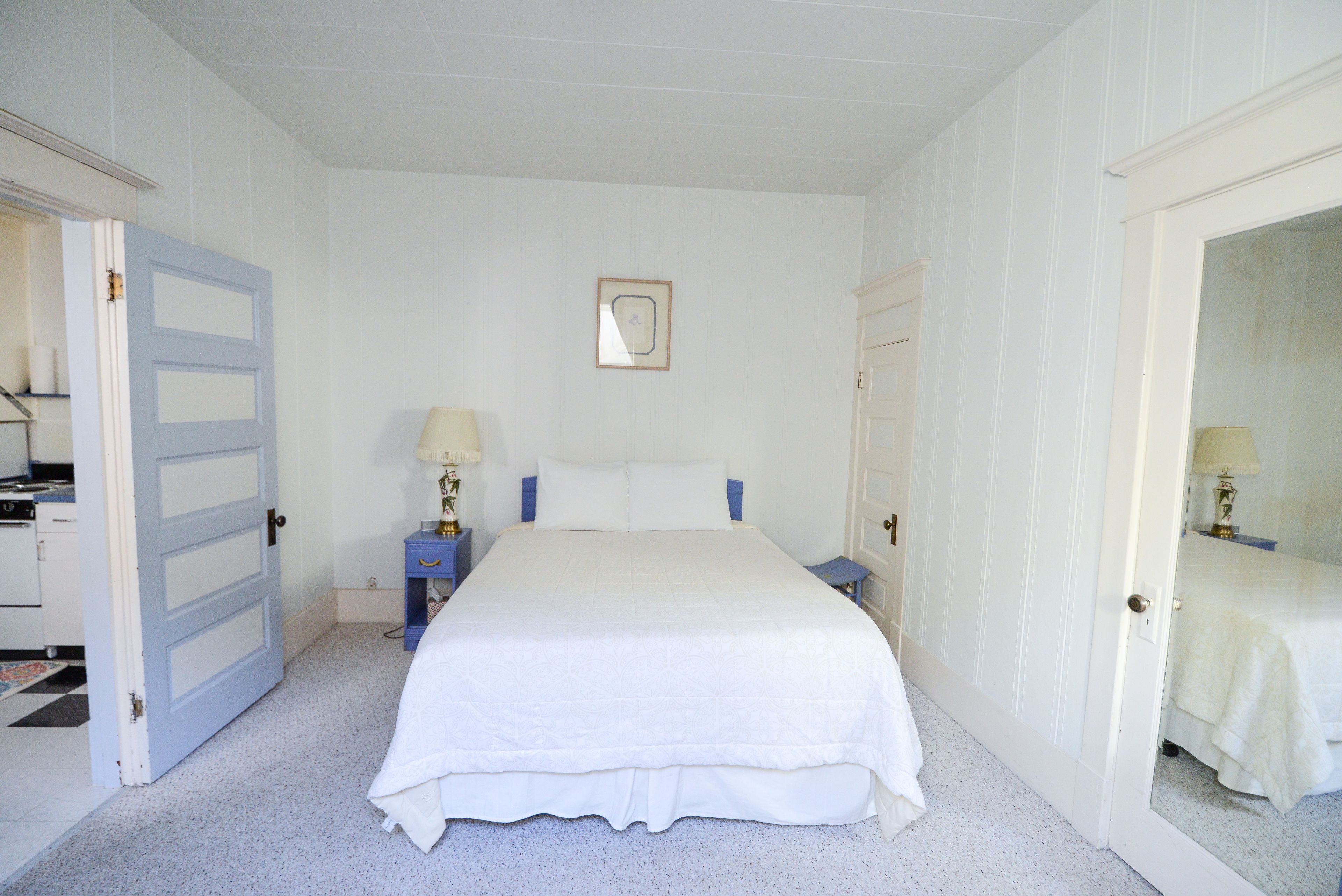 Une chambre avec un grand lit, deux tables de chevet, une commode, un tableau encadré au mur et une porte donnant sur une autre pièce.