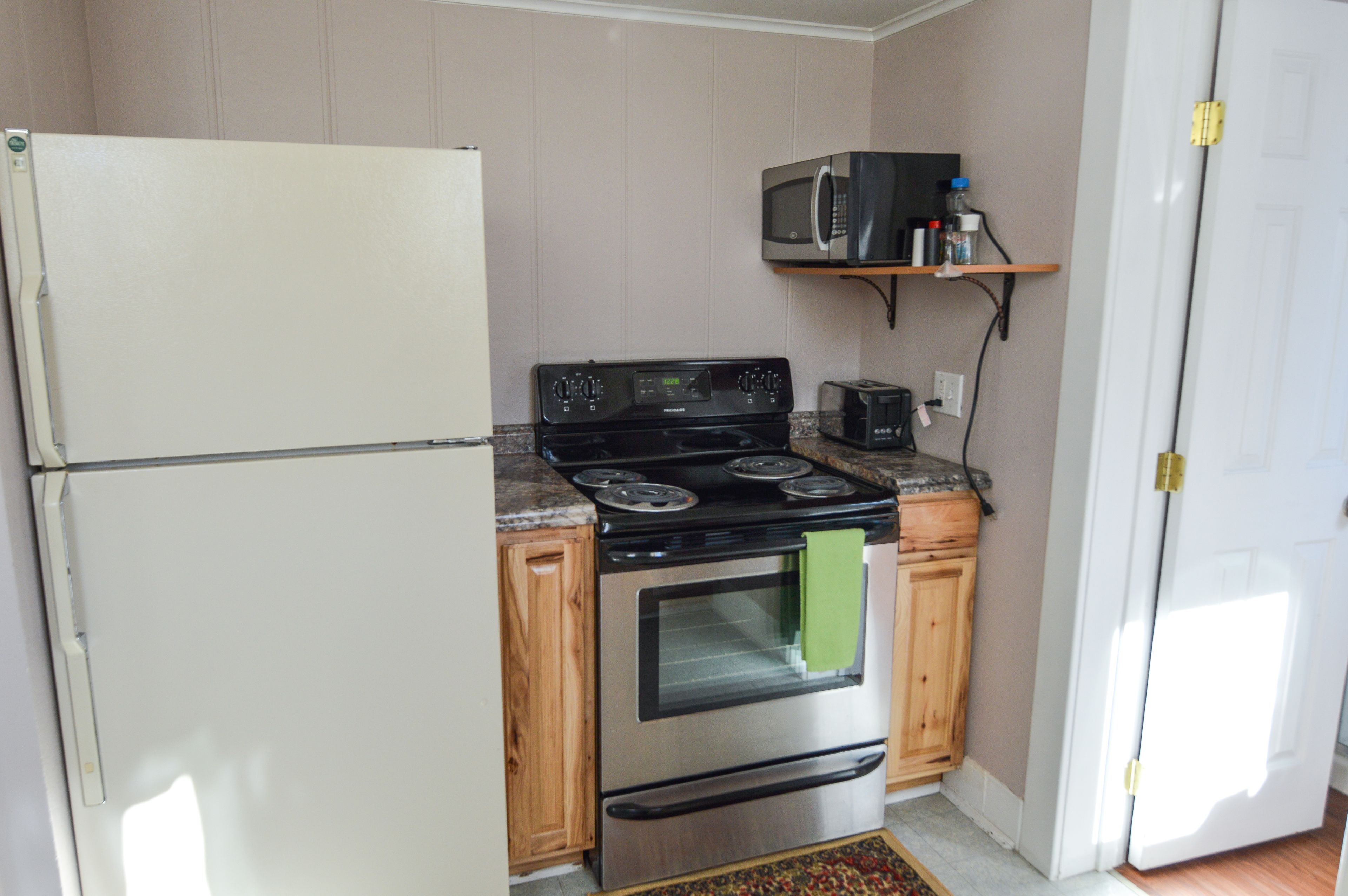 Eine kleine Küche mit Kühlschrank, Herd, Backofen, Mikrowelle und einem Holzregal über dem Herd.
