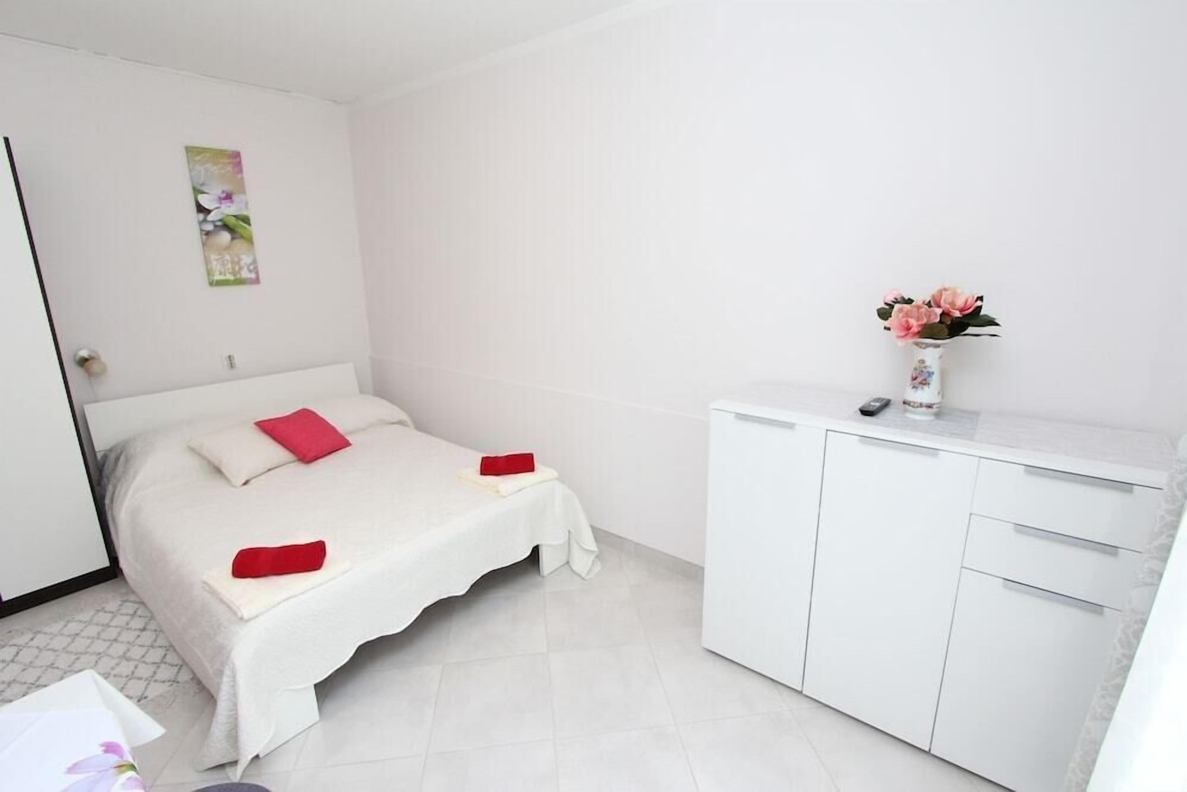 Een nette, witte slaapkamer met een bed, een nachtkastje, een kast en een vaas met bloemen.