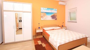 Un dormitorio con cama, armario, mesita de noche, lámpara, un cuadro de una playa y ventana.