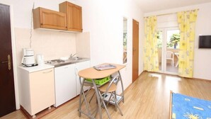 Una cocina compacta con un lavamanos, una cafetera y una mesita. Hay un armario de madera sobre el lavamanos y un caja verde para almacenamiento en el suelo. La habitación tiene piso de madera y una puerta que da a un área exterior con cortinas amarillas.