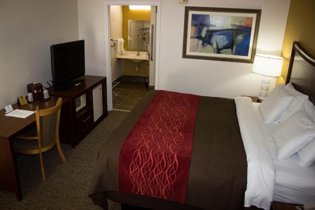 A hotel room with a bed, a desk with a television, a chair, a painting on the wall, and a bathroom visible through an open door.