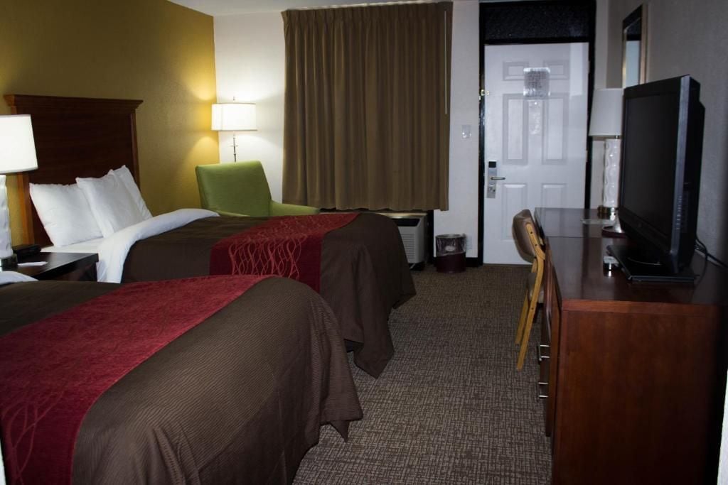Ein Hotelzimmer mit zwei Betten, einem Schreibtisch mit Fernseher, einem Sessel und einer Tür.