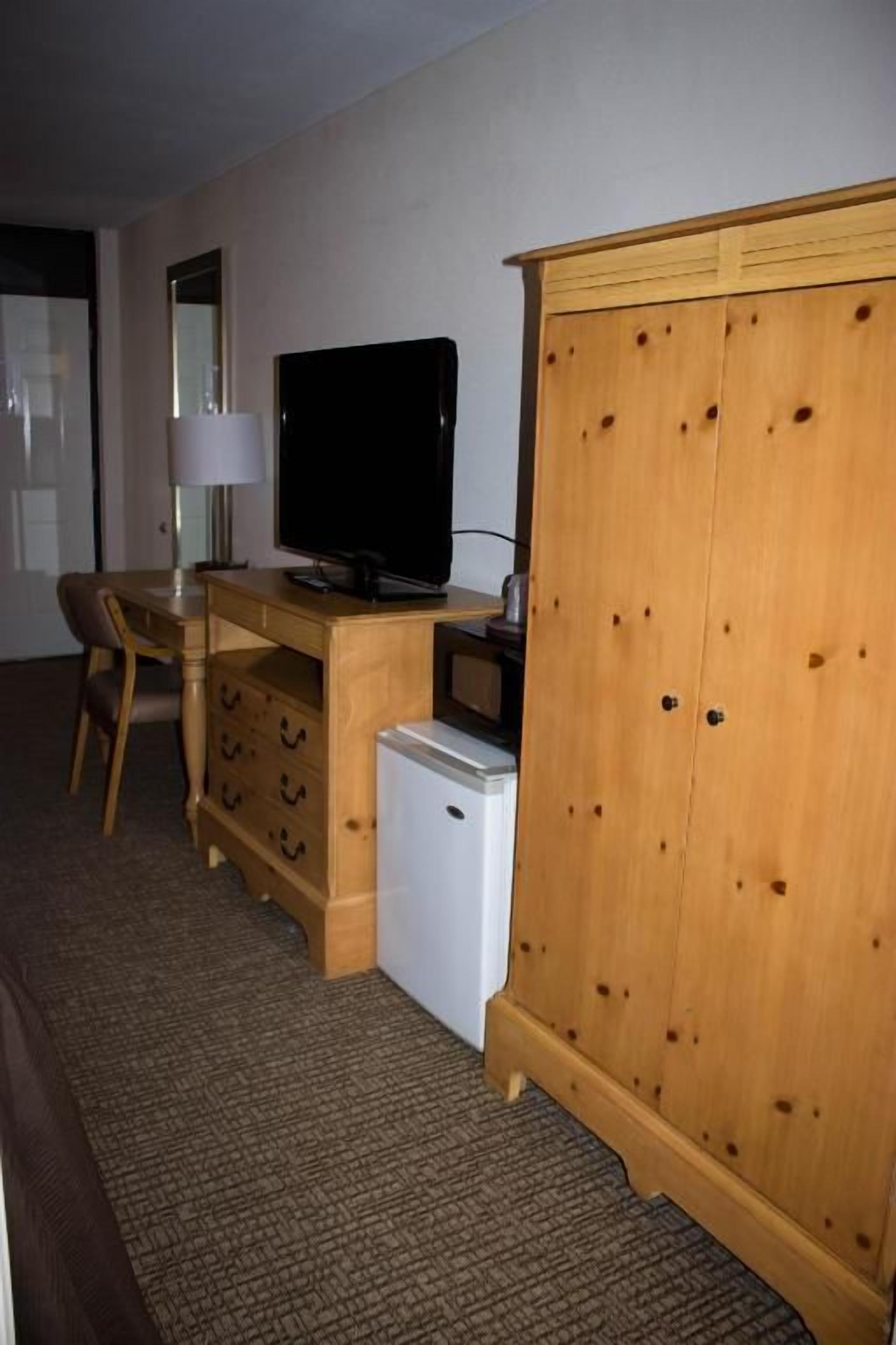A room with a wooden desk, a television, a small refrigerator, and a wardrobe.