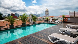 A rooftop swimming pool with lounge chairs, potted plants, and a view of a city skyline including a prominent building with a dome.