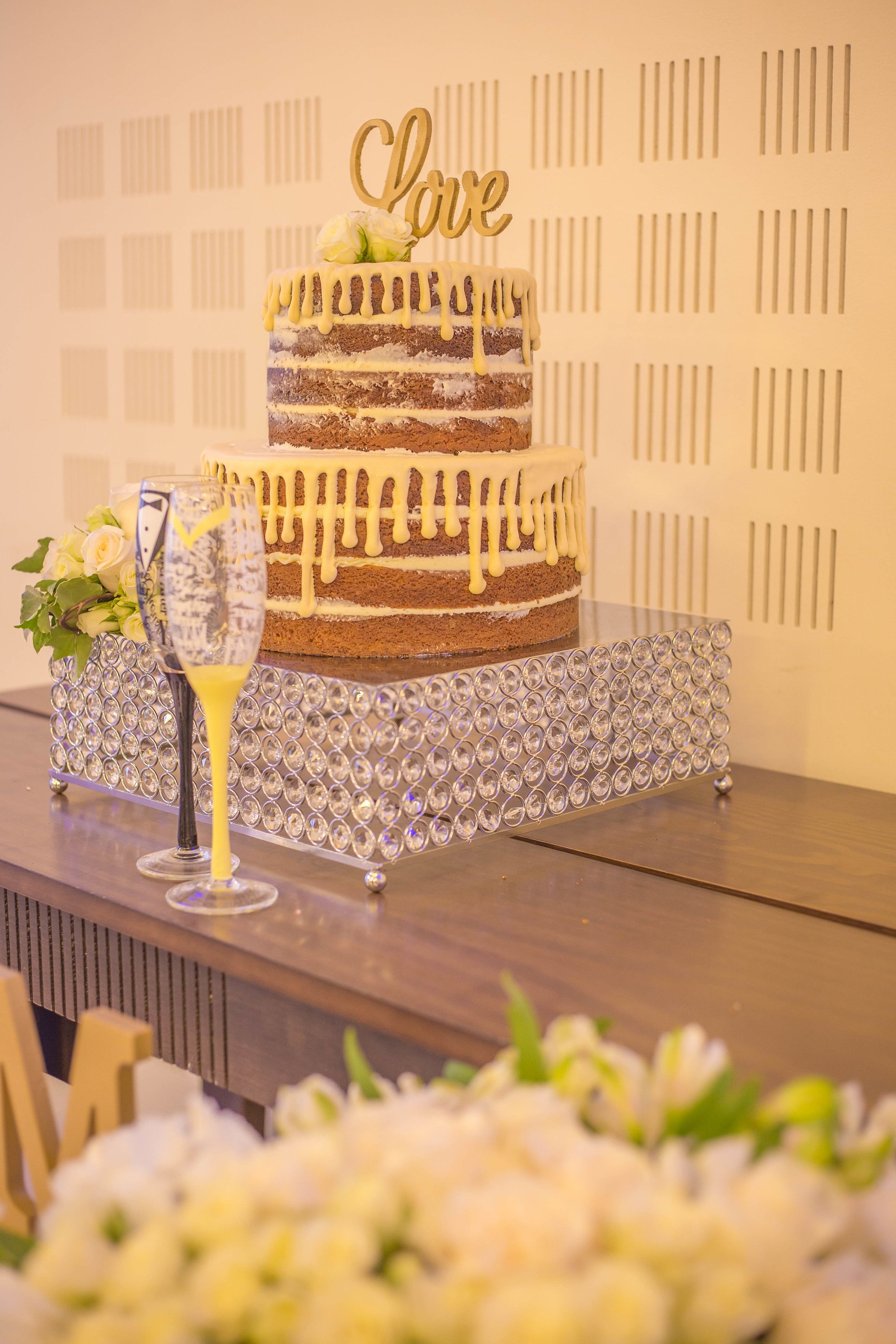 Un gâteau à plusieurs étages, surmonté d’un décor en forme de cœur, posé sur un présentoir décoratif, avec une coupe de champagne à côté.