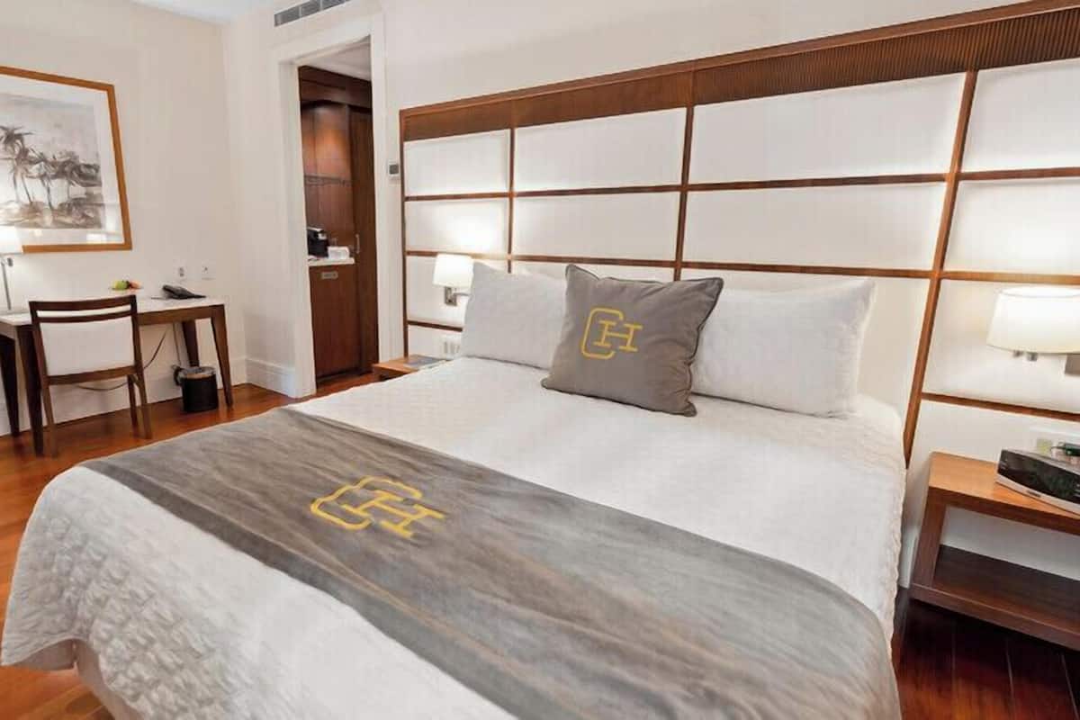 Une chambre d’hôtel avec un grand lit, un bureau et une chaise. Il y a un tableau au mur et une tête de lit en bois.