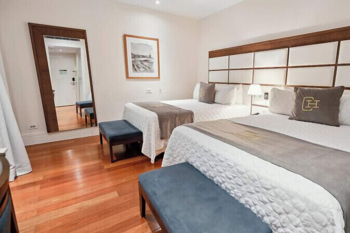 Une chambre d’hôtel avec deux lits, un banc, un miroir et un tableau encadré au mur.