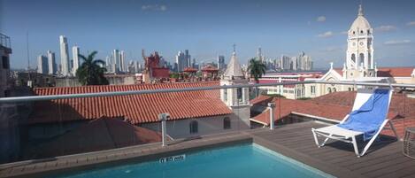 A rooftop pool with a blue chair, a city skyline in the background, and a bell tower.