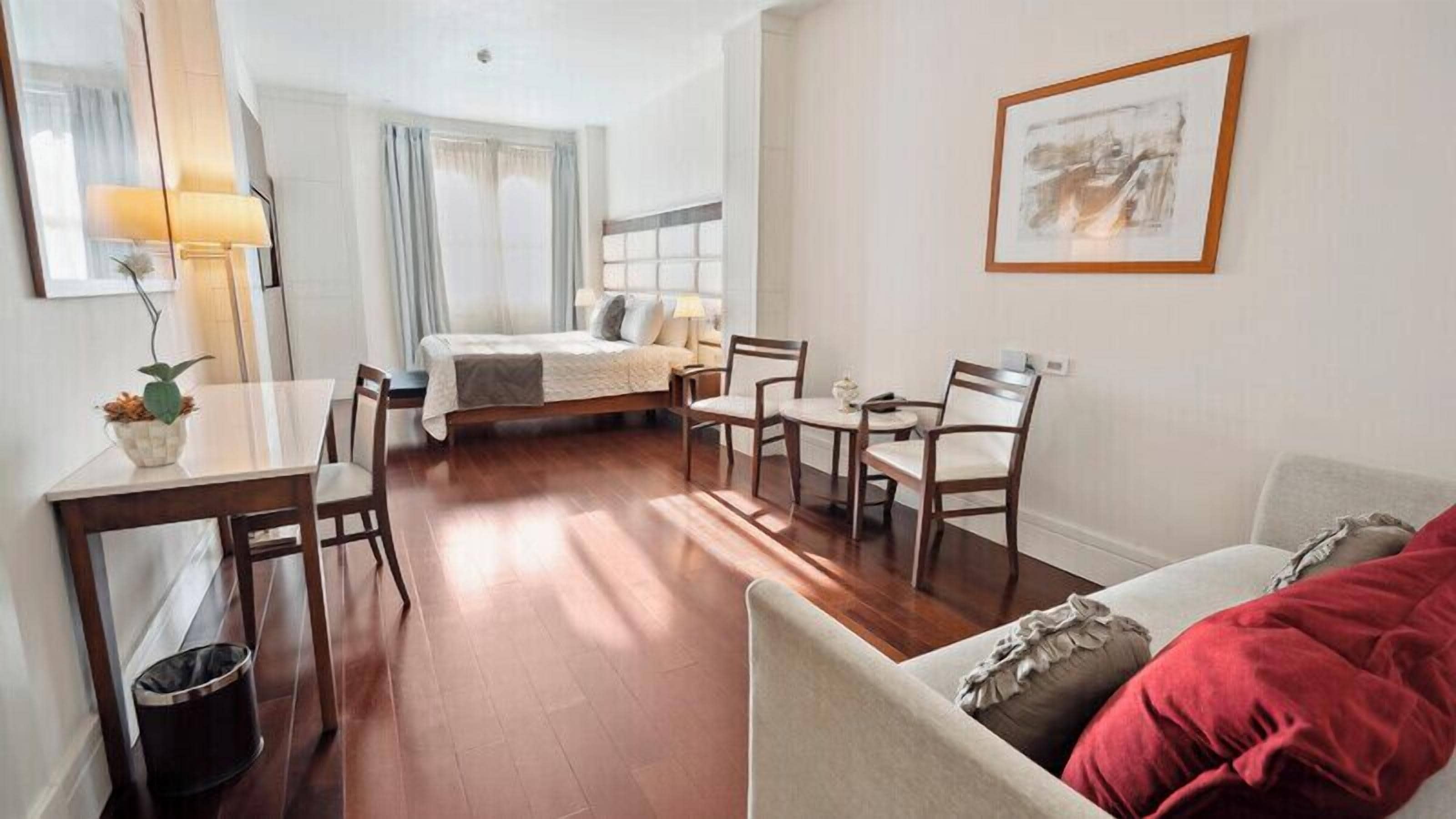 A compact hotel room with a bed, a dining table with chairs, a small sofa, a trash bin, and a framed picture on the wall.
