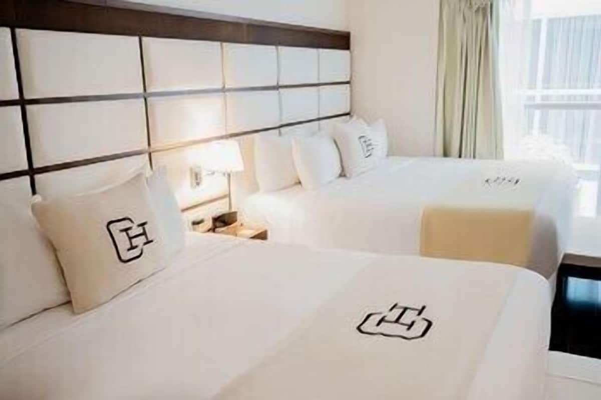 Une chambre d’hôtel avec deux lits, une tête de lit à motifs géométriques, une table de chevet avec une lampe et une fenêtre avec des rideaux.