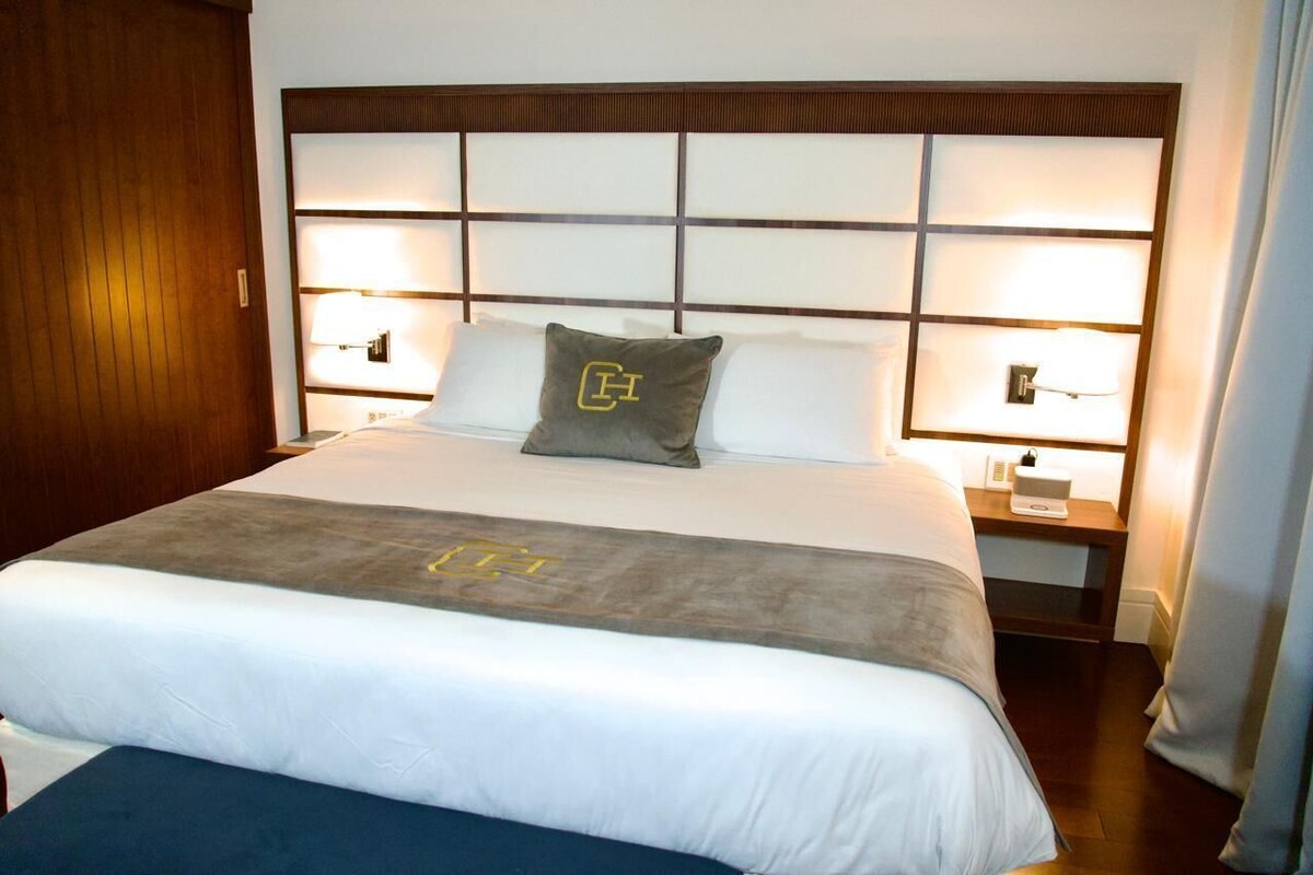 A hotel room with a large bed, two bedside tables, a headboard with a grid pattern, and a nightstand with a lamp.