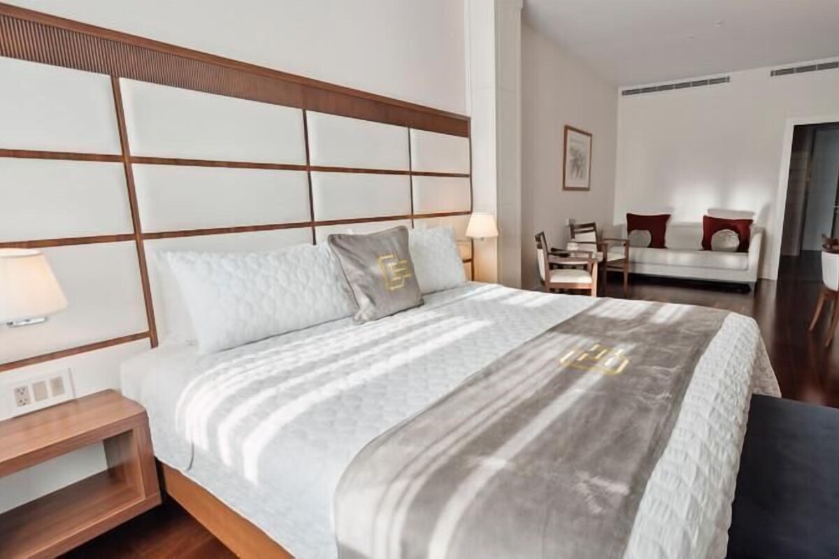A hotel room with a large bed, bedside table, lamp, and a view of the dining area.