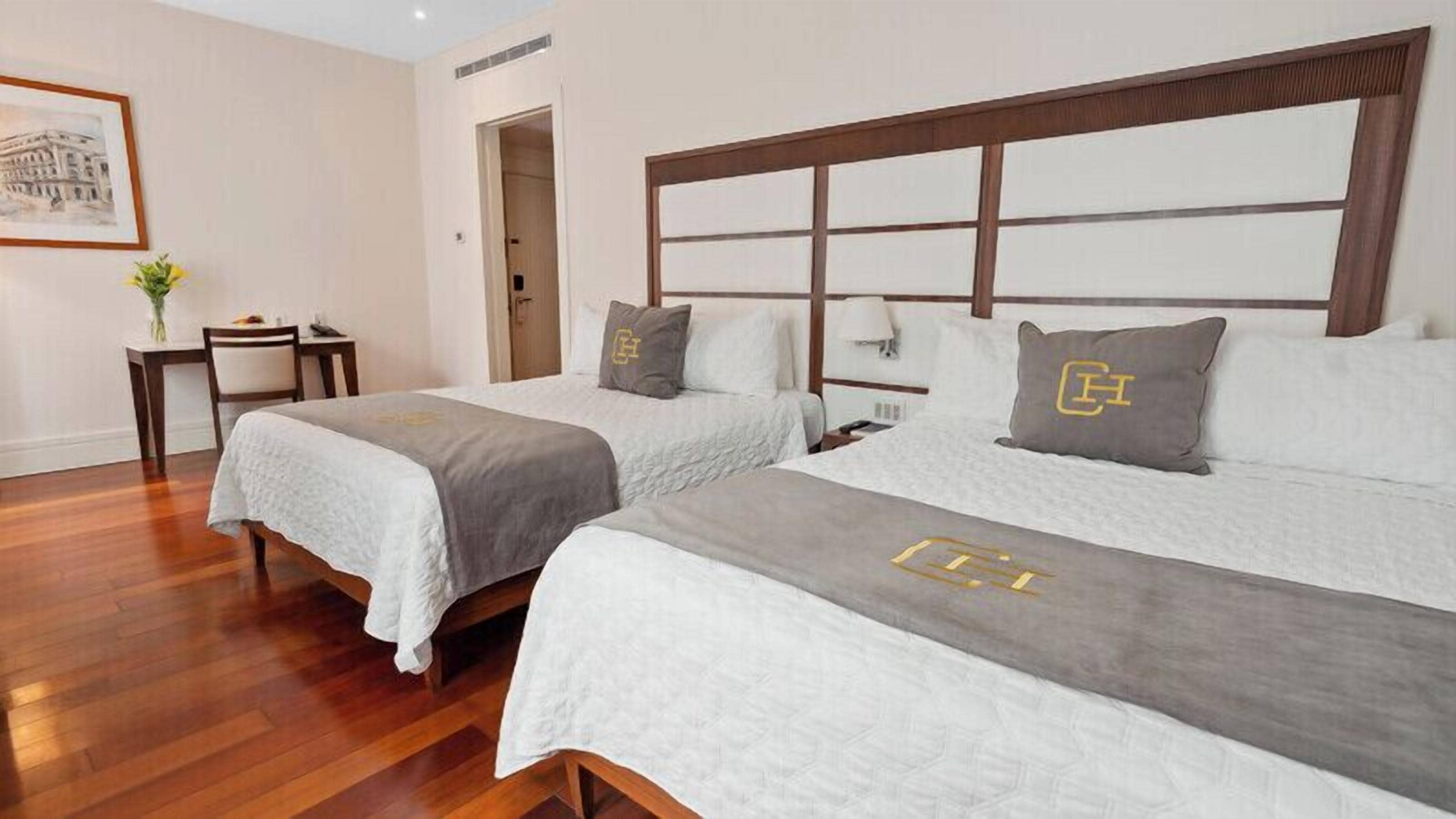 Habitación de hotel con dos camas, cabecero de madera, una mesita con un florero y un cuadro en la pared.