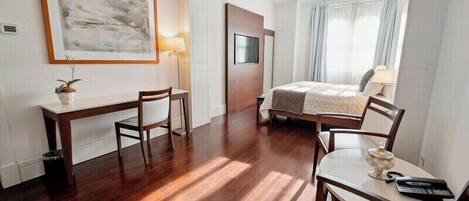 Habitación de hotel con piso de madera, una cama con cubrecama blanco, un escritorio con silla, una mesita de noche con lámpara y un cuadro enmarcado en la pared.