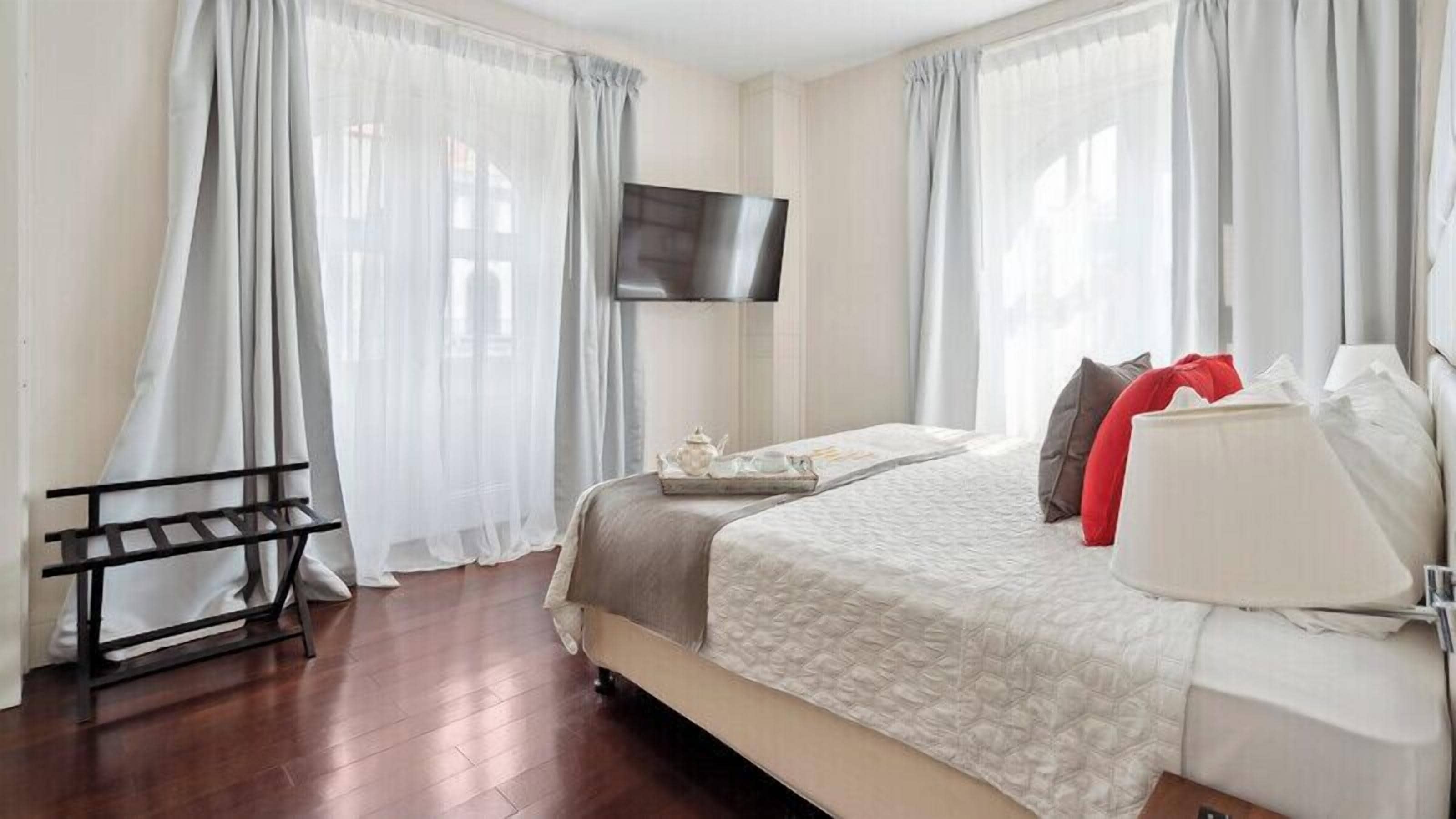 A hotel room with a bed, a TV mounted on the wall, and a window with sheer curtains.