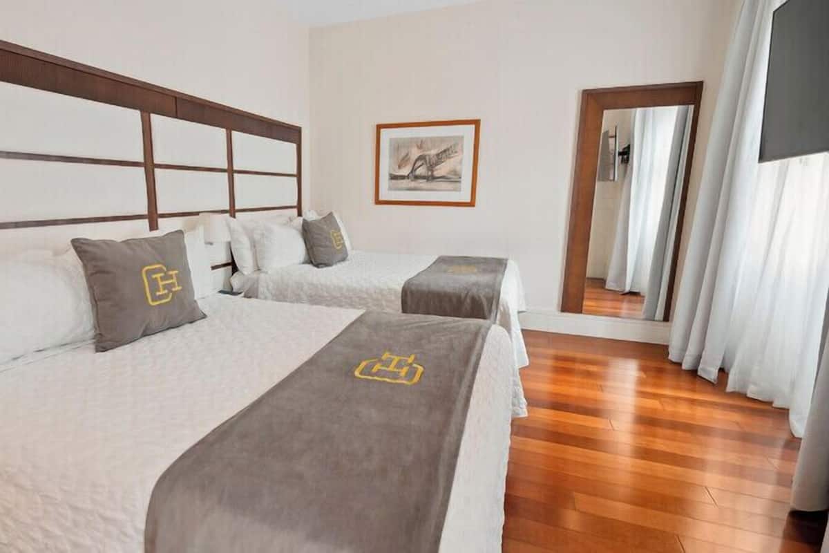 Une chambre d’hôtel avec deux lits, une tête de lit en bois, un tableau encadré au mur et un miroir qui reflète la pièce.