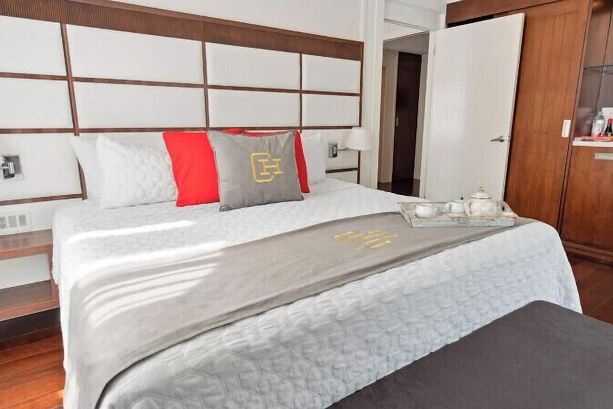 Un lit bien fait, avec une couette grise et blanche, un coussin rouge et un coussin gris orné d’un logo. Sur le lit, on aperçoit un plateau avec une théière et des tasses.