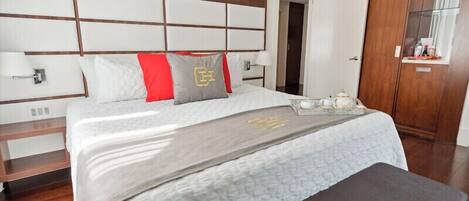 A neatly made bed with a grey and white duvet, a red throw pillow, and a grey pillow with a logo. There is a tray with a teapot and cups on the bed.
