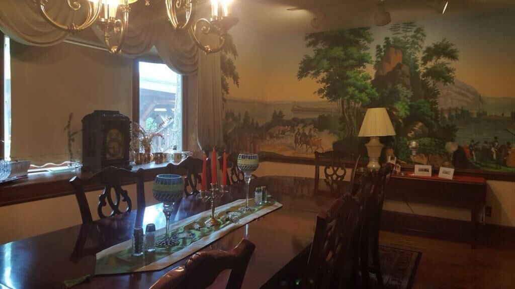 Et spiseområde med et langt bord dækket til et måltid, omgivet af stole. En lysekrone hænger fra loftet, og en mural der viser en historisk scene er synlig på væggen bag bordet.