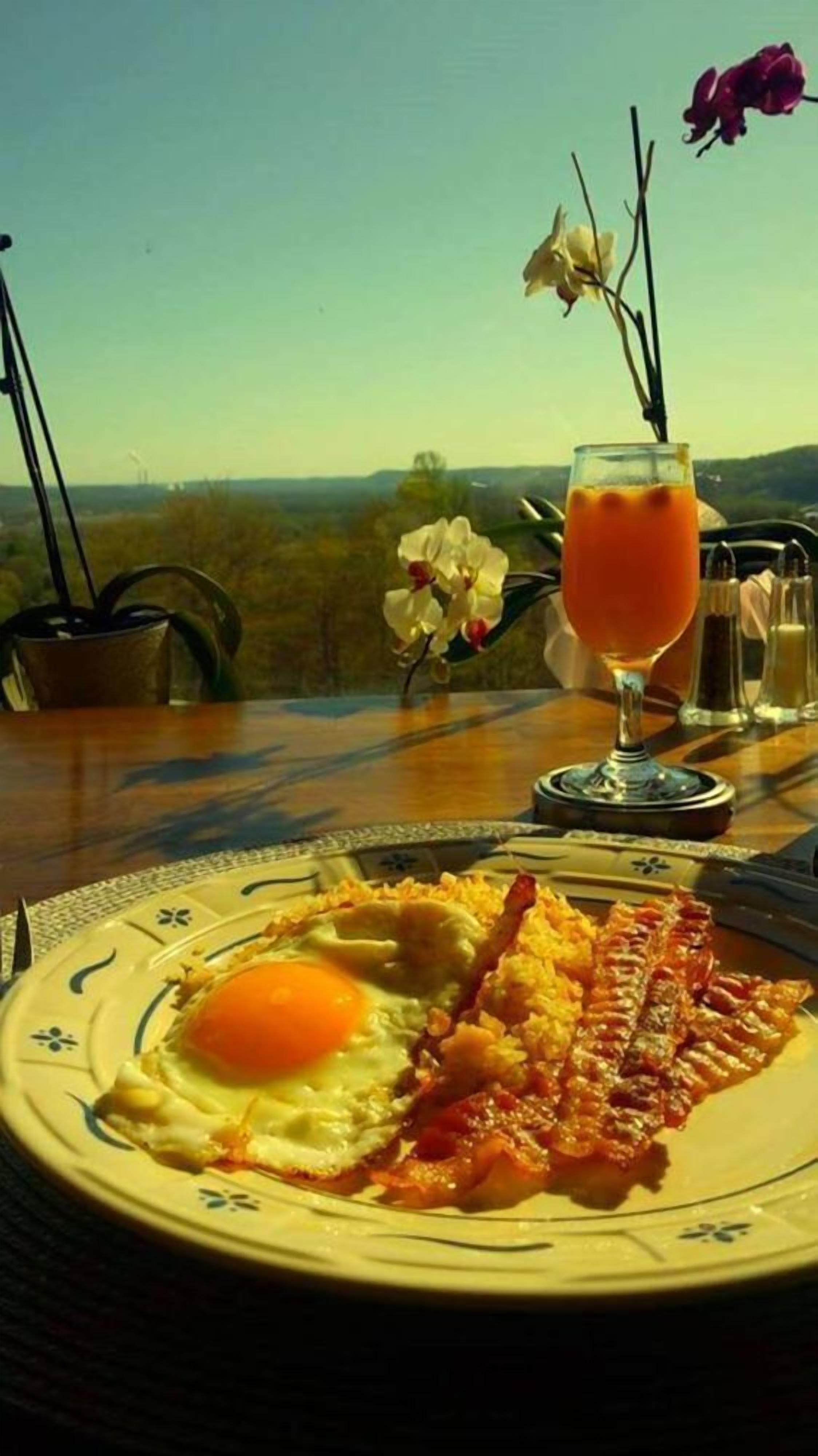 Une assiette de petit-déjeuner avec un œuf et des lardons, un verre de jus d’orange et une vue sur un paysage fleuri sous un ciel dégagé.