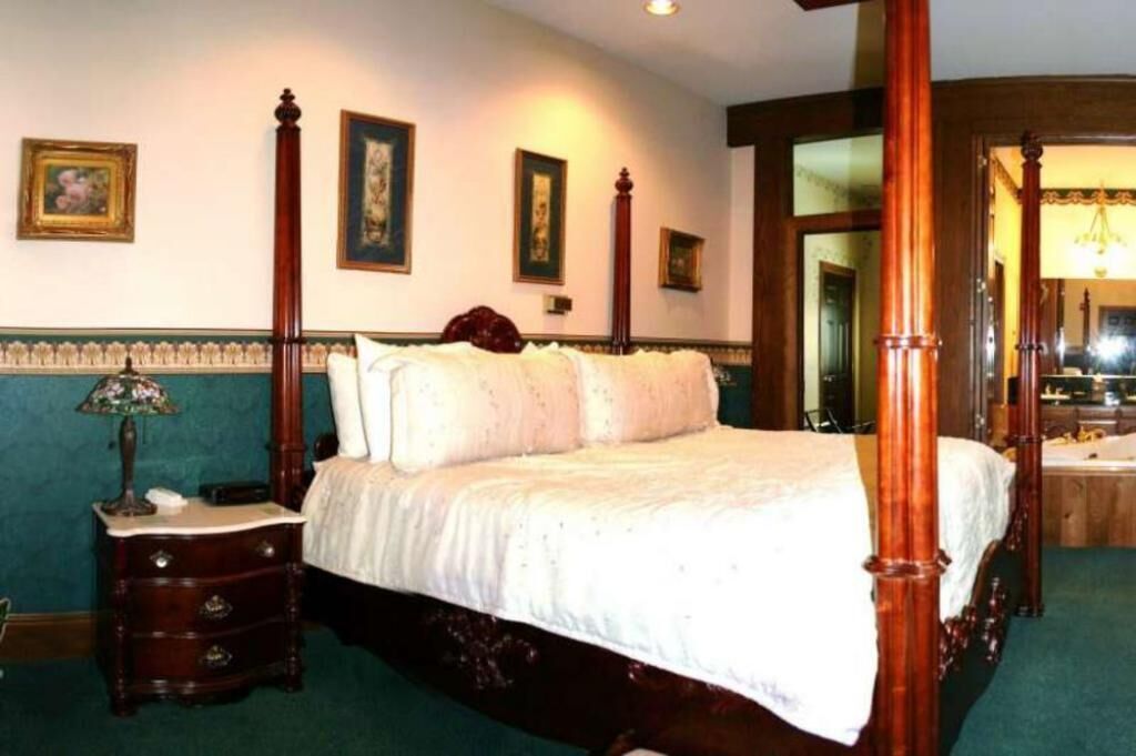 Un lit à baldaquin avec une structure en bois, une tête de lit rembourrée de tissu vert et un couvre-lit blanc. Il y a une table de chevet avec une lampe et un miroir.