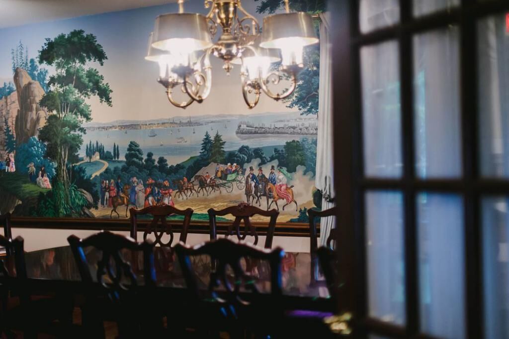 A dining area with a mural depicting a historical scene, a chandelier, and wooden chairs.