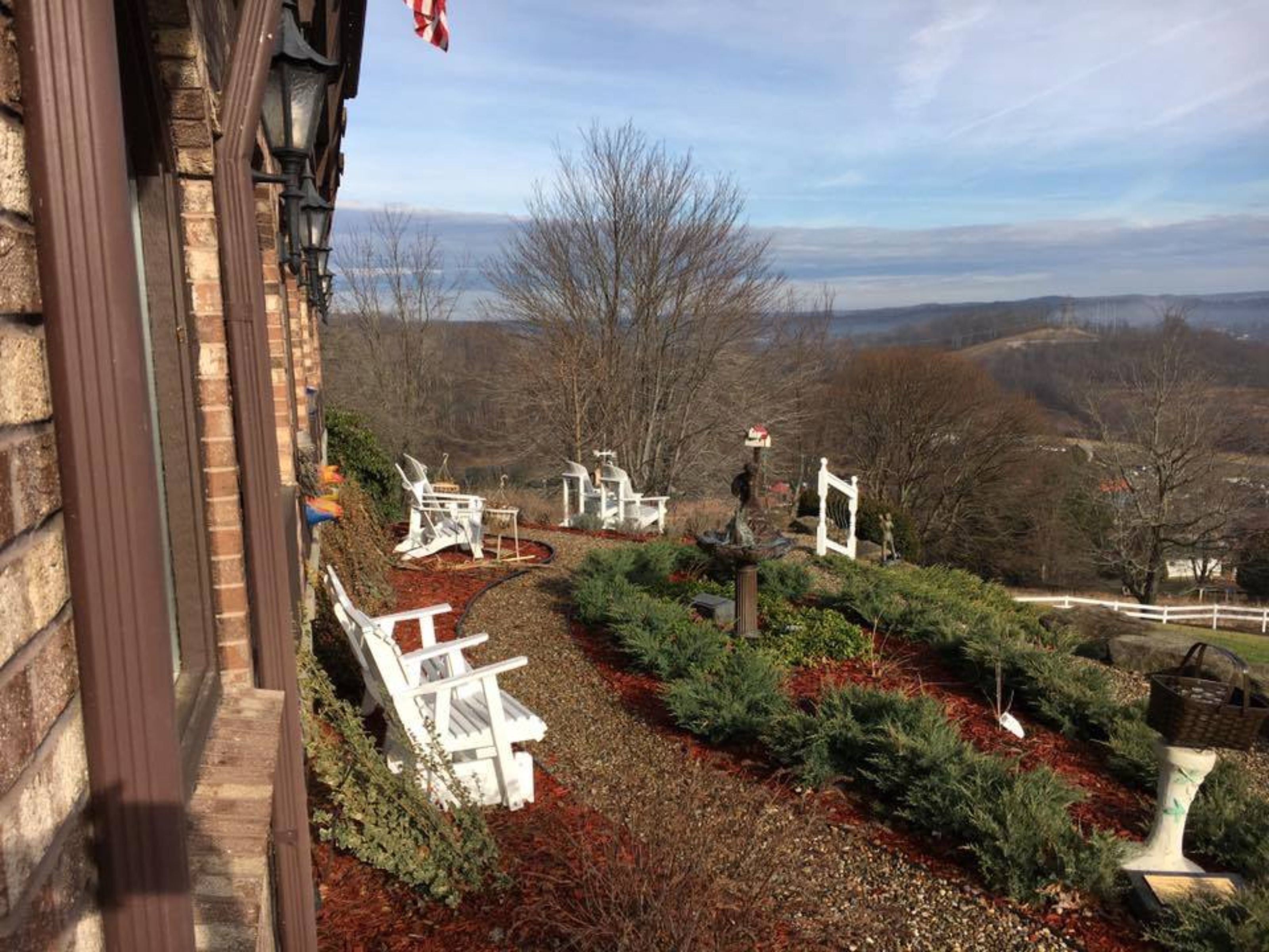 Un jardin avec des chaises blanches et une statue, entouré d’arbres et offrant une vue sur des collines lointaines.