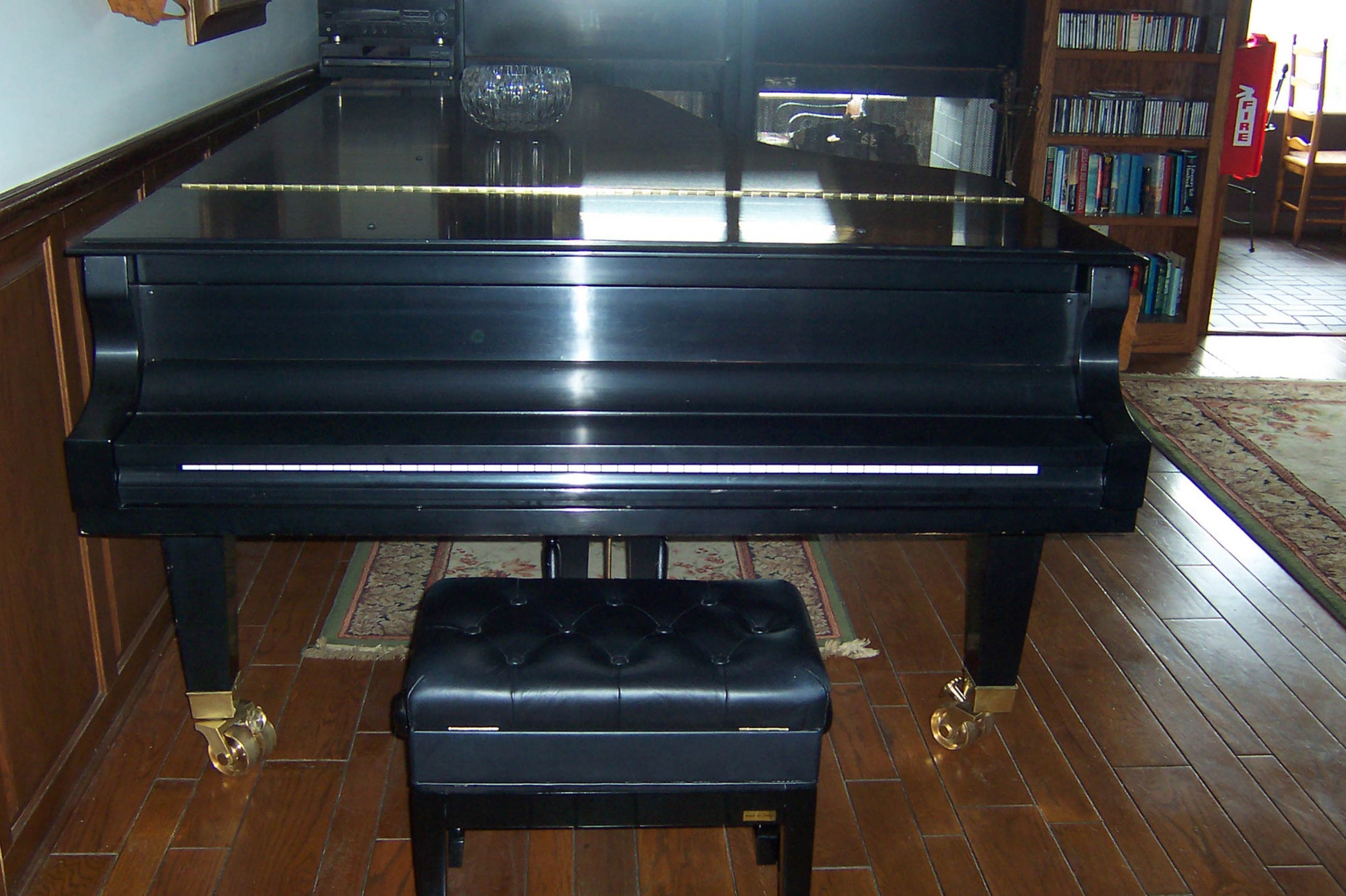 Un piano de cola en una habitación con piso de madera y una estantería al fondo.