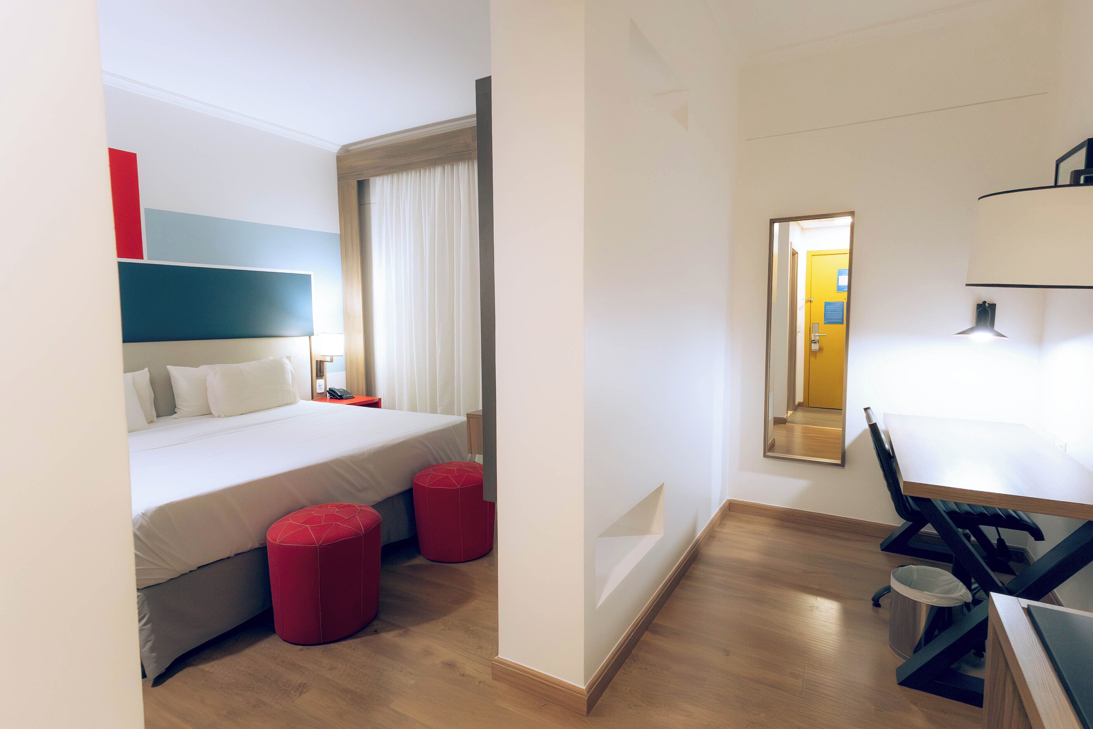 Habitación de hotel con una cama, dos otomanos rojos, un escritorio con una silla y un espejo en la pared.