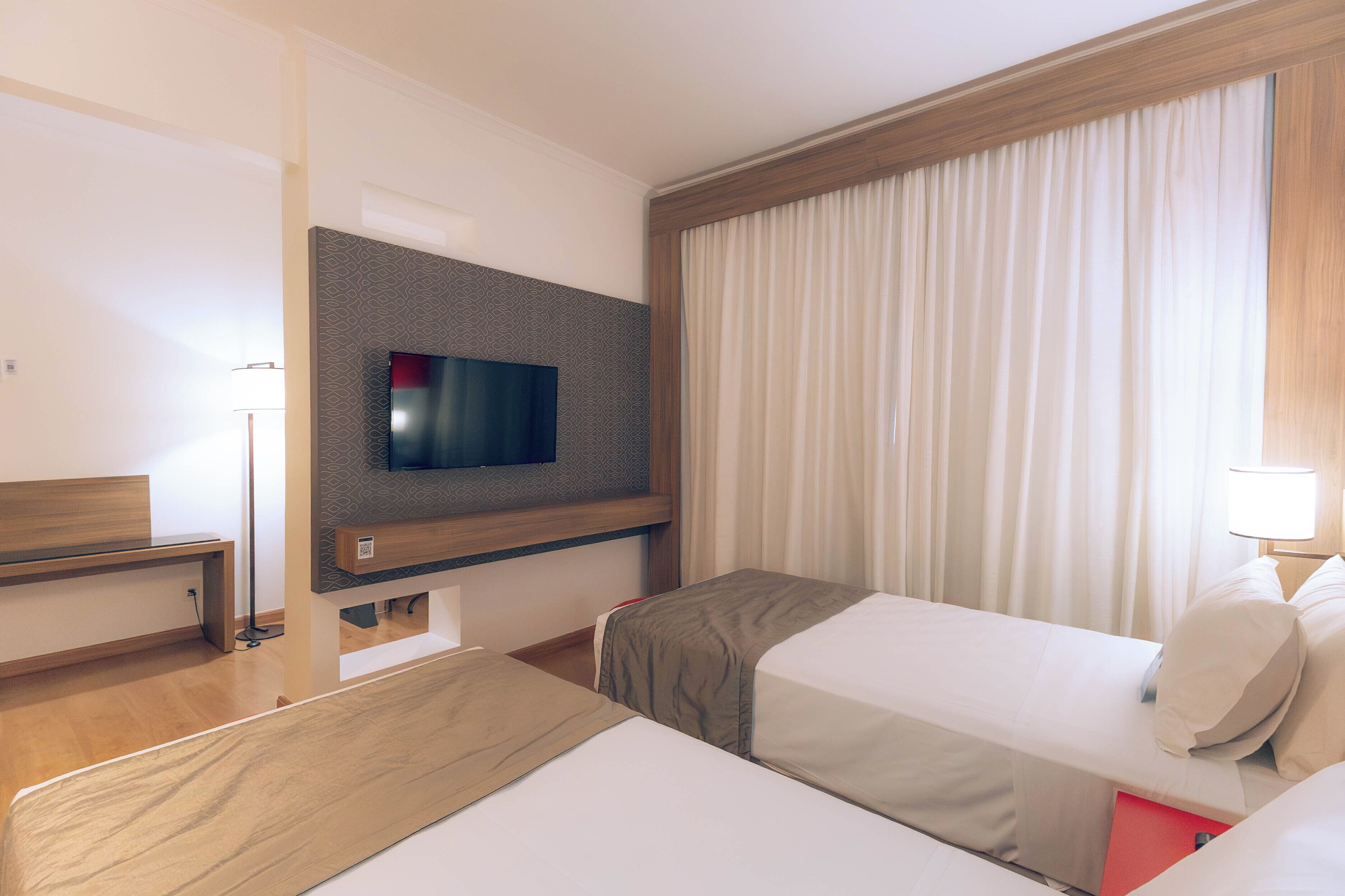 A hotel room with two beds, a TV mounted on the wall, and a large window with curtains.