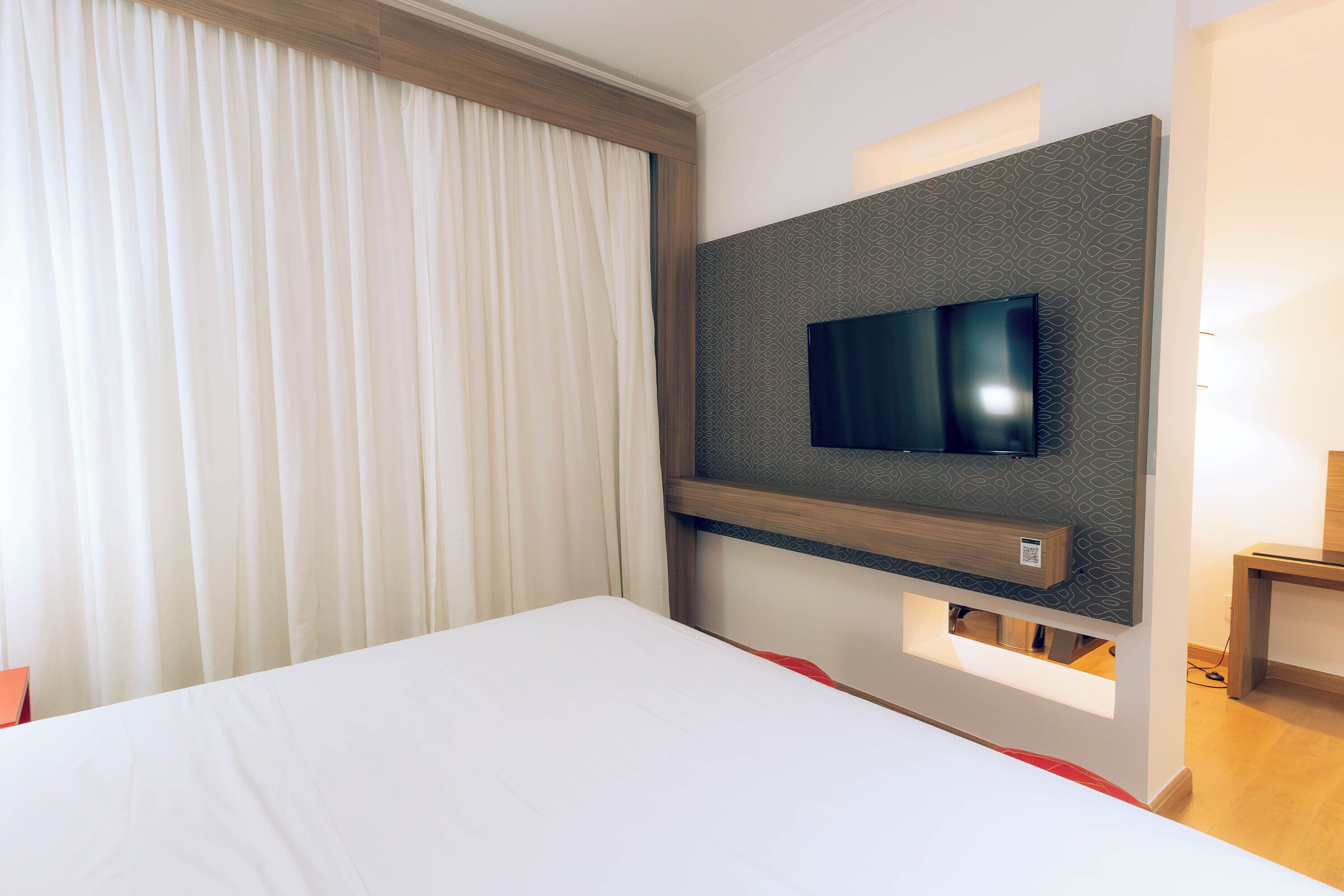 A hotel room with a large bed, a mounted flat-screen TV, and a wooden nightstand.
