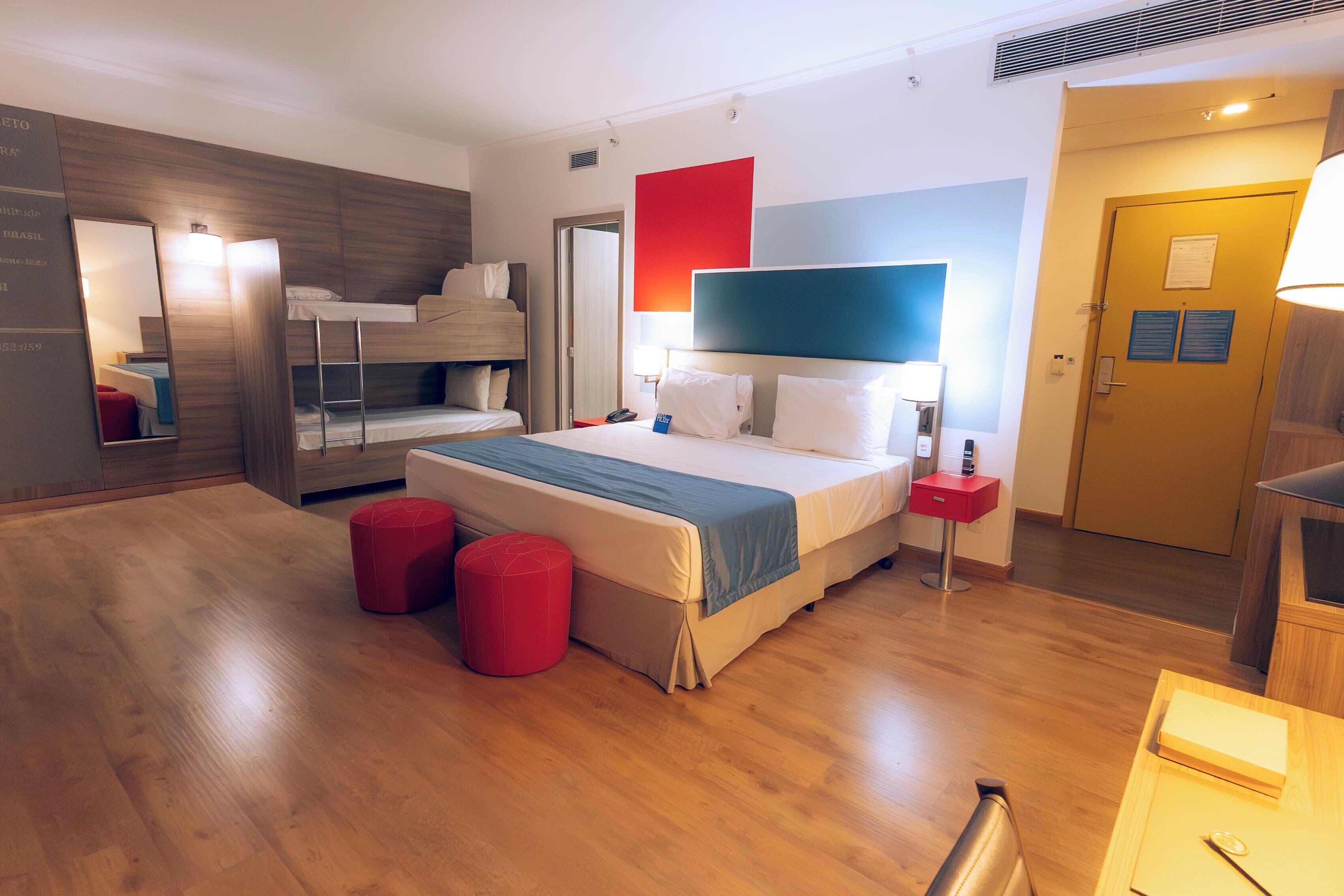 Una habitación de hotel moderna con una litera, una cama grande con cabecera azul, piso de madera y un reposapiés rojo.