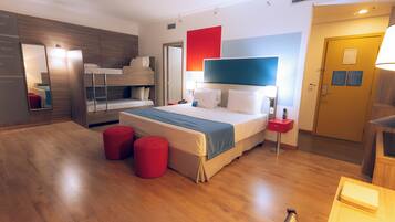 Una habitación de hotel moderna con una litera, una cama grande con cabecera azul, piso de madera y un reposapiés rojo.