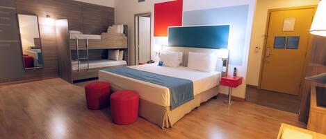 A modern hotel room with a bunk bed, a large bed with a blue headboard, wooden flooring, and a red ottoman.