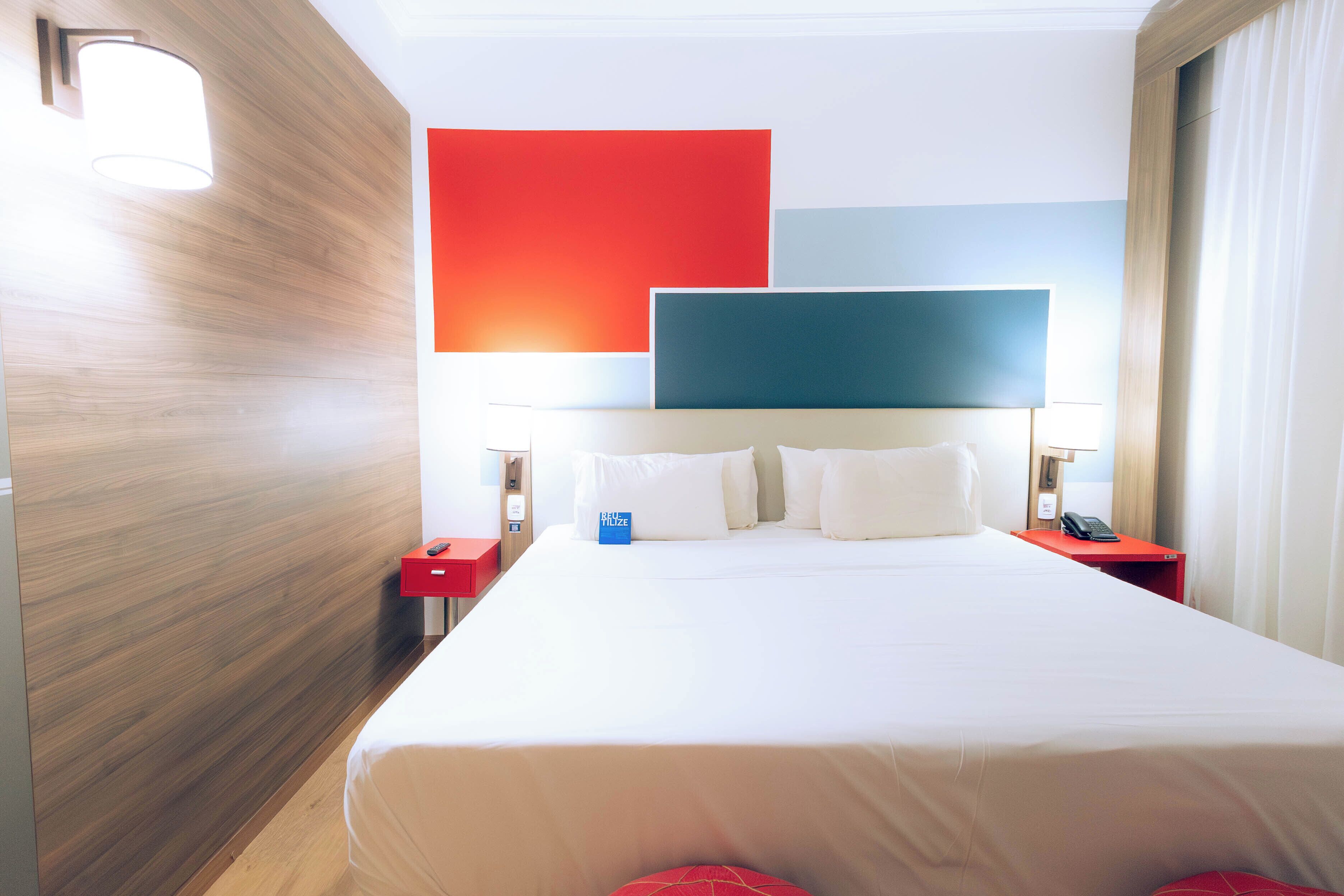 A hotel room with a large bed, a red nightstand, a blue pillow, and a red pillow.